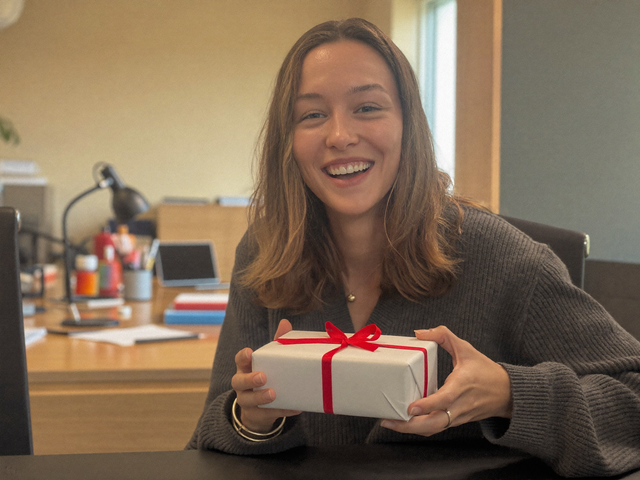 Lächelnde Frau hält ein verpacktes Geschenk mit roter Schleife an ihrem Büro-Schreibtisch