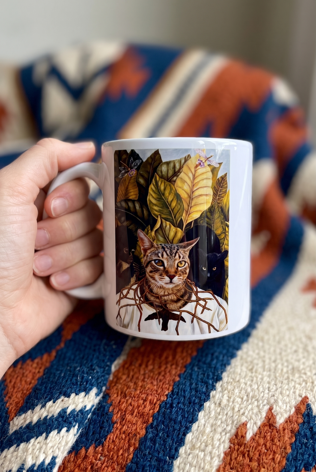 Bunte Tasse mit Katze in der Hand ueber einer gewebten Decke