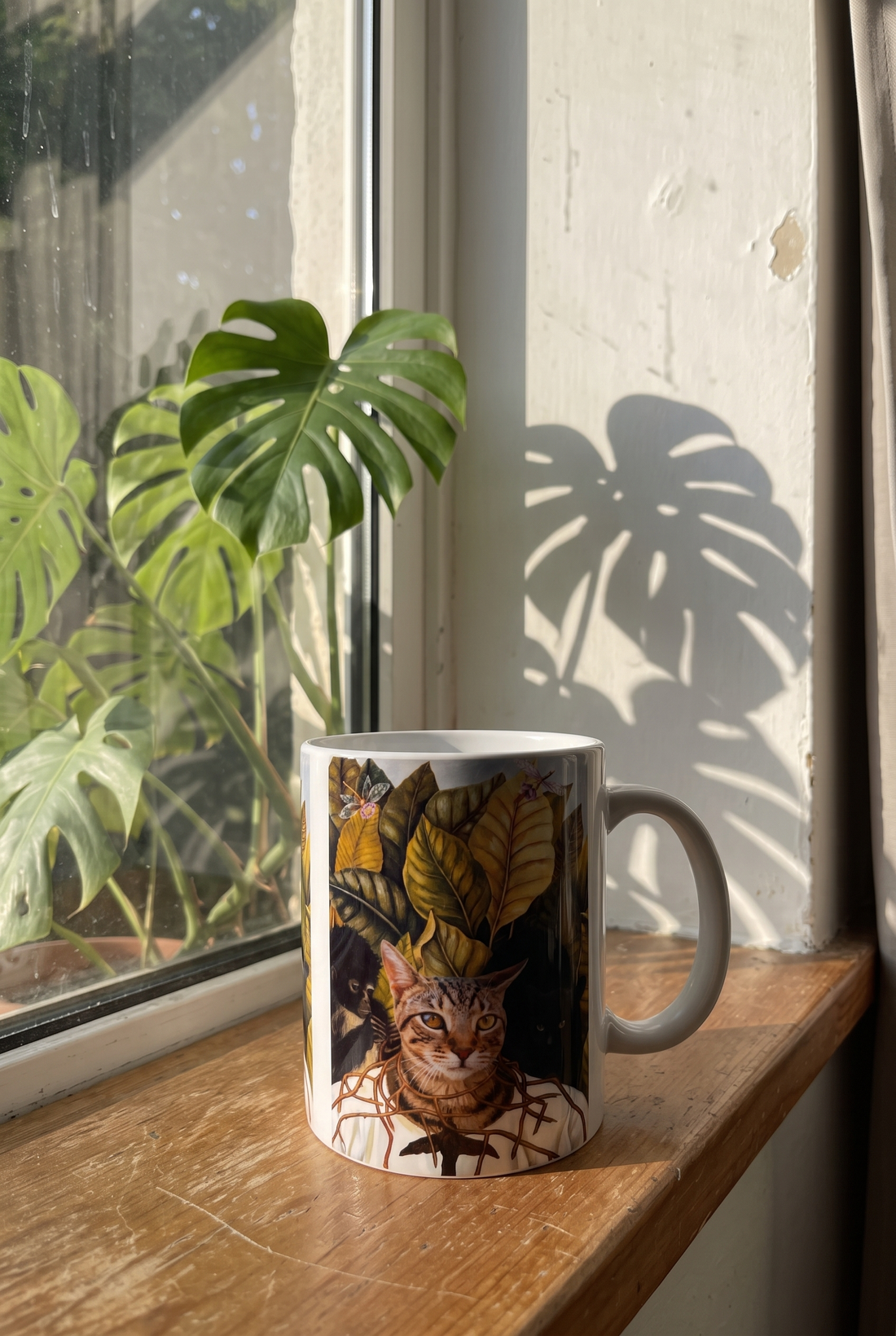 Bunte Tasse mit Katze auf einer Fensterbank vor einer Monstera